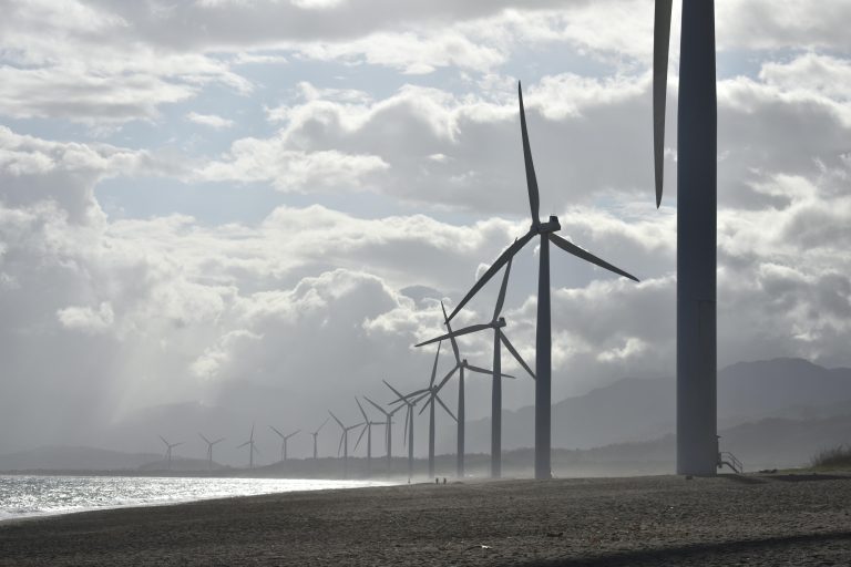 A row of wind turbines along a cloudy beach shoreline in Vigan City, Philippines.