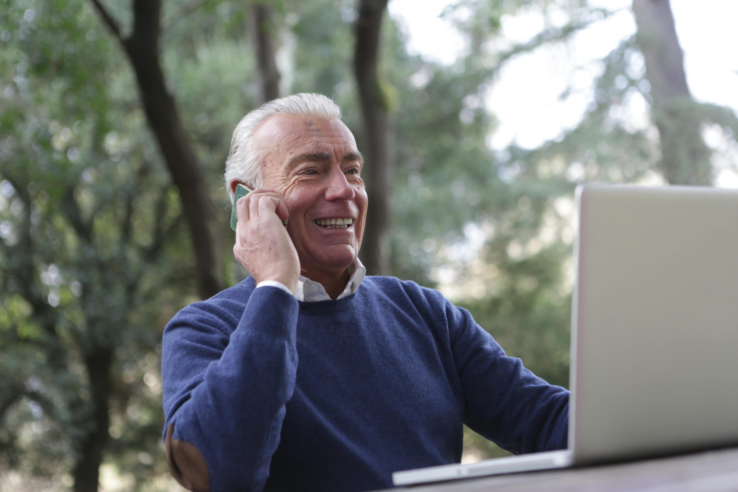 Elderly man joyfully using a laptop and phone outdoors, embracing technology.