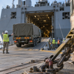 A secured cargo crate and lashing chain in focus while logistics crews guide a supply truck onto a transport ship.